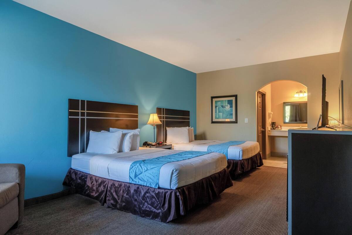 A hotel room with two beds, a desk, a chair, a mirror, and a bathroom visible through an archway.