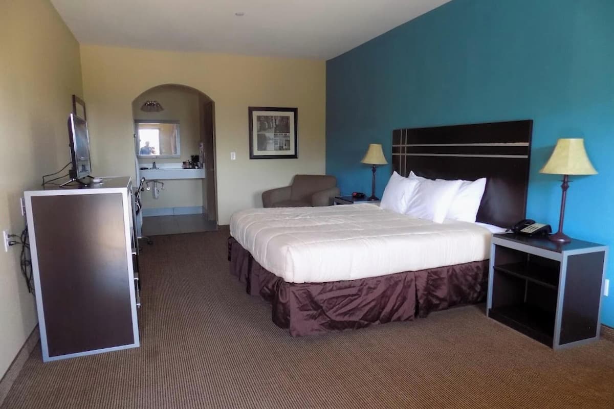 A hotel room with a bed, bedside tables, a desk, a chair, a desk lamp, a telephone, a mirror, and a bathroom visible through an archway.