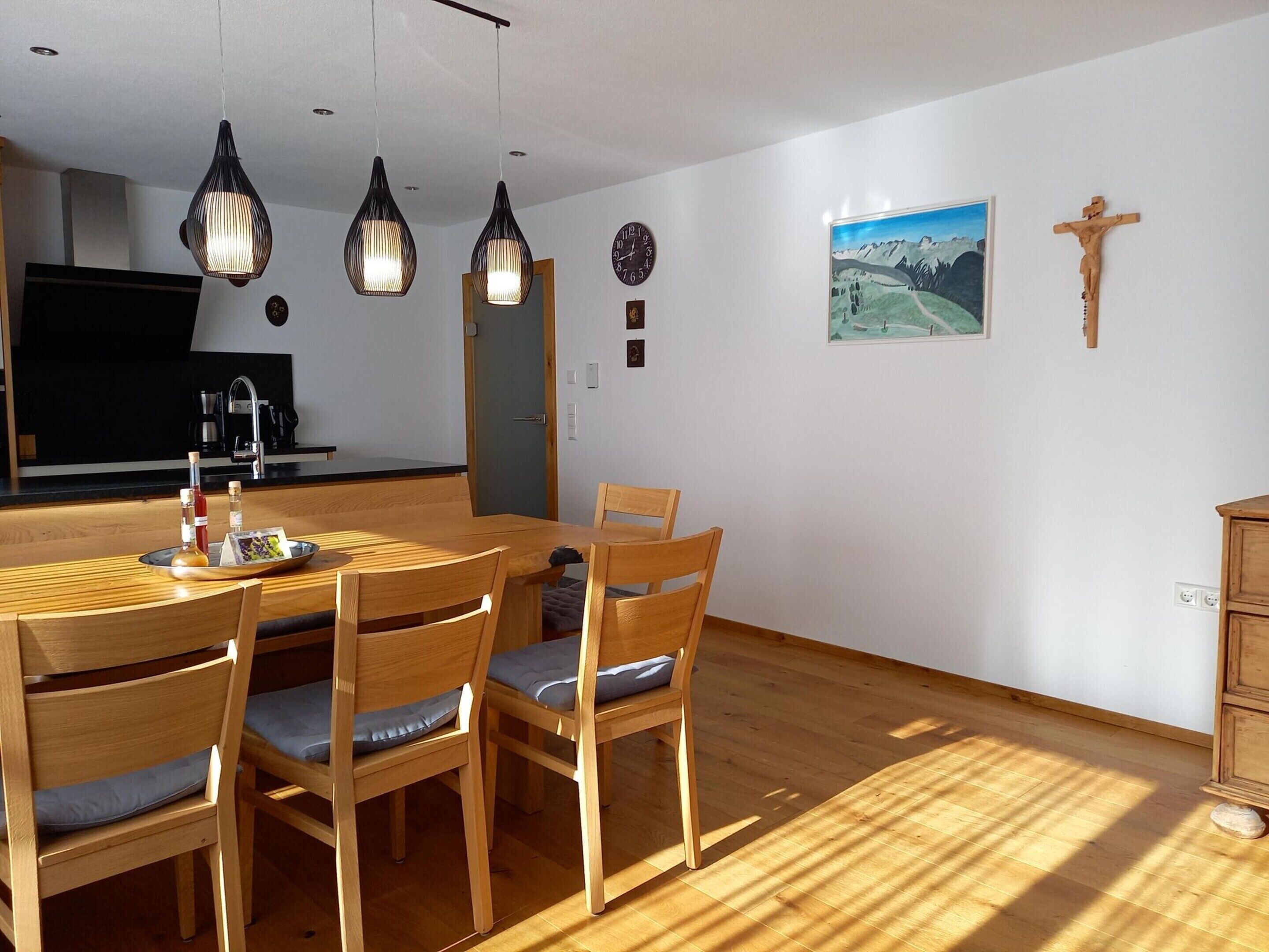 Un coin repas avec une table et des chaises en bois, des luminaires suspendus, une cuisine en arrière-plan, un tableau représentant un paysage et un crucifix en bois accroché au mur.