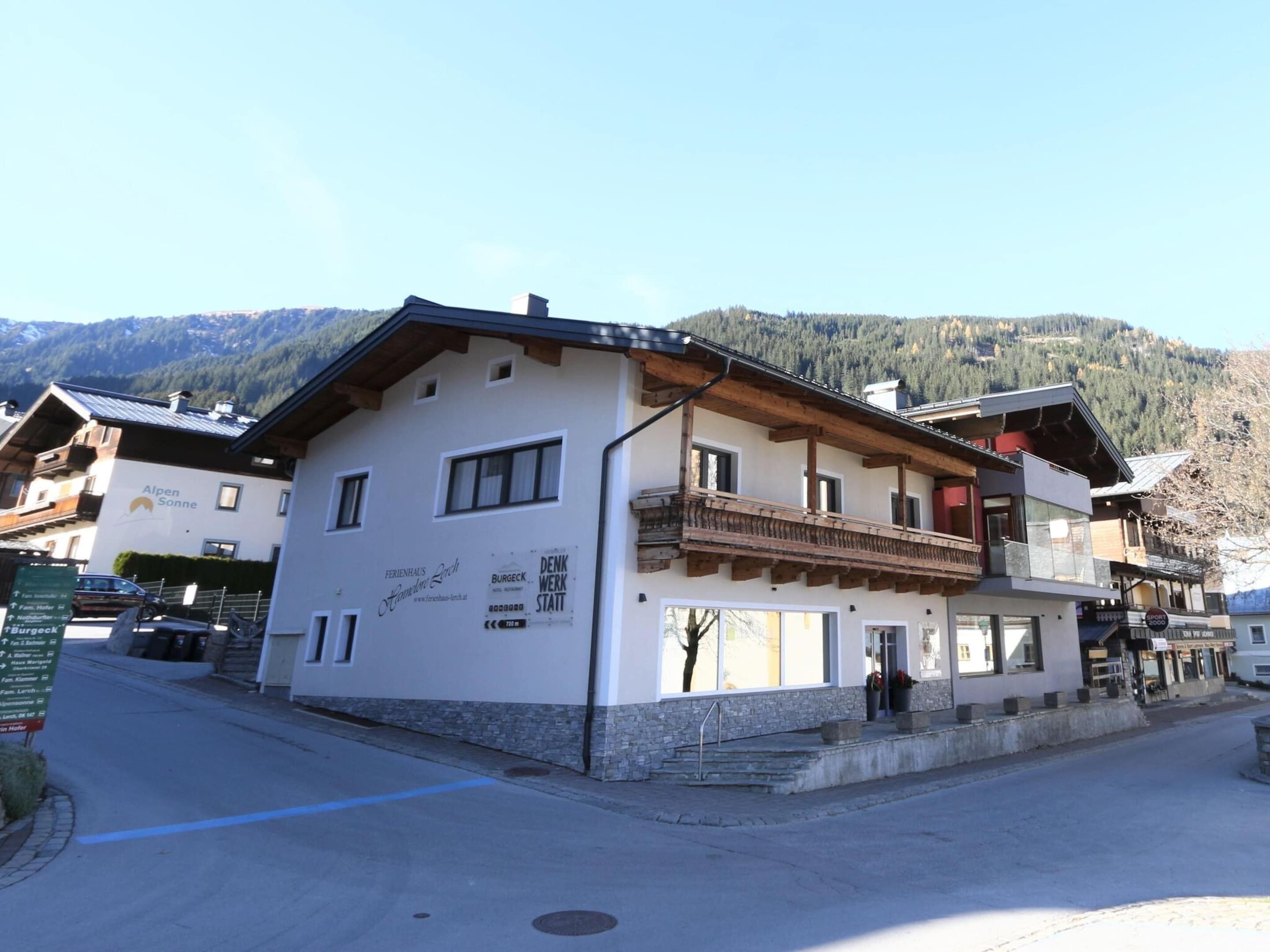 Ein zweigeschossiges Gebäude mit Balkon, einem Schild mit der Aufschrift 'Alpen Sonne' und Bergelandschaft im Hintergrund.