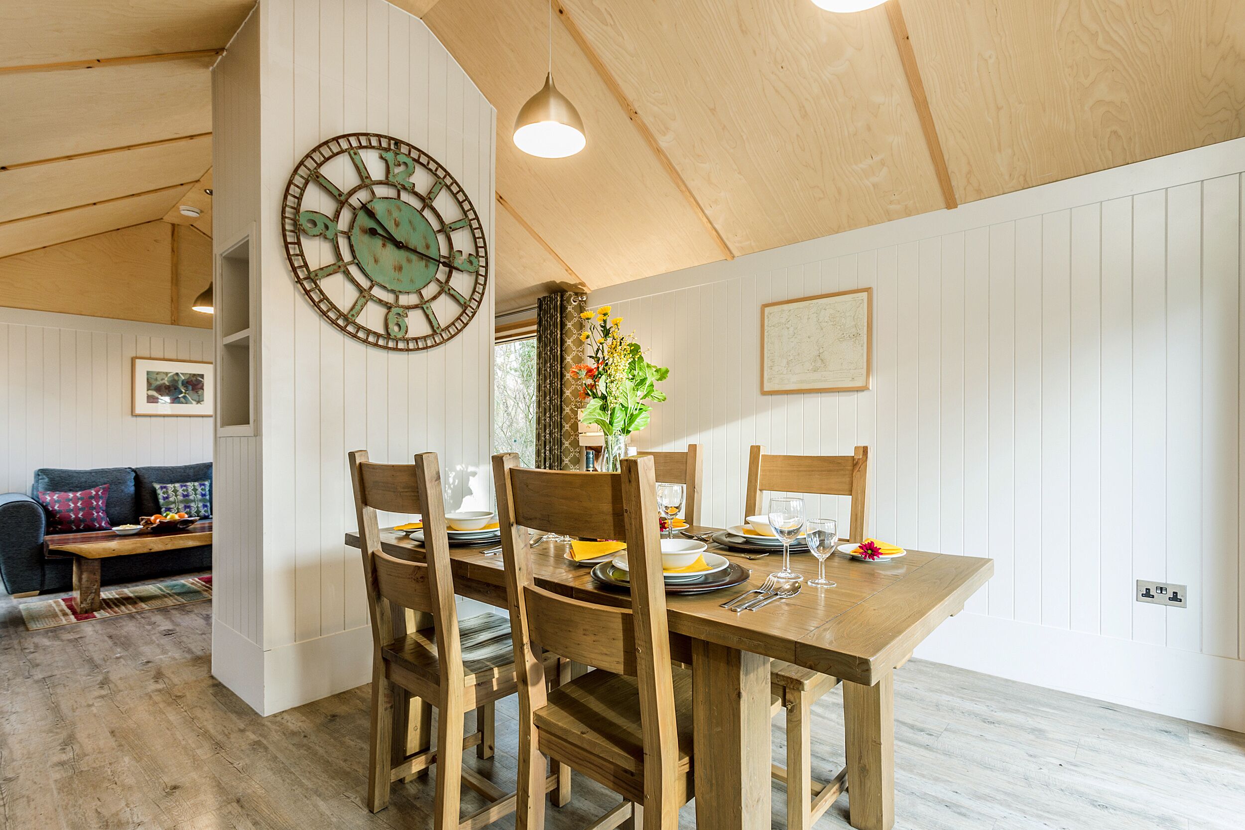 A dining area with a wooden table set for four, surrounded by wooden chairs. A large wall clock is mounted on the wall above the table.