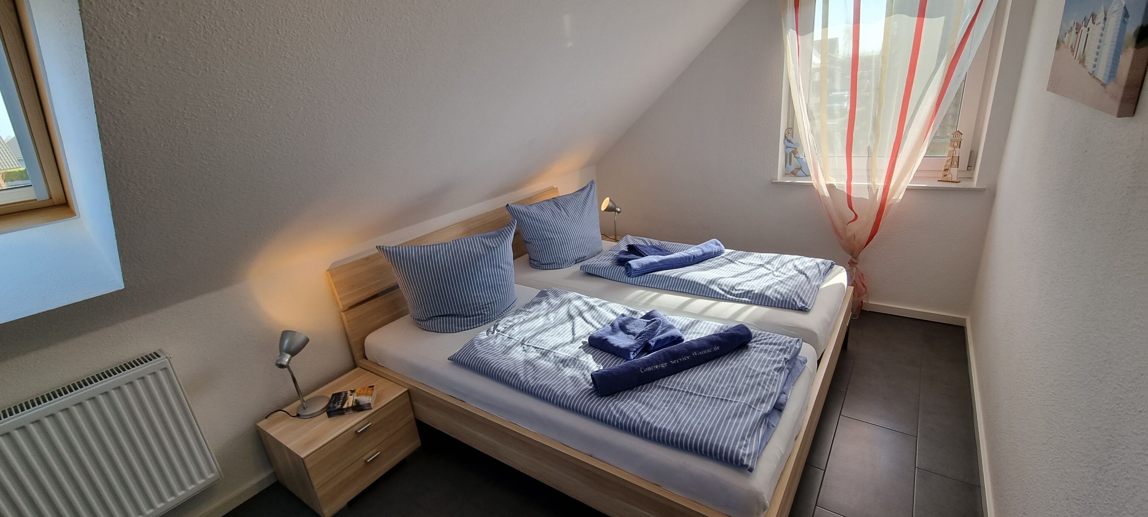 A bedroom with two beds, a bedside table with a lamp, a radiator, and a window with a view of buildings.