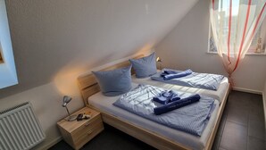 A bedroom with two beds, a bedside table with a lamp, a radiator, and a window with a view of buildings.