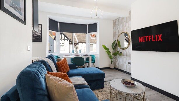 A modern living room with a blue sofa, a dining area with a table and chairs, a large window, and a wall-mounted TV labeled "NETFLIX".