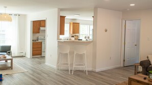 A modern kitchen with wooden cabinets, a bar area with two white bar stools, and a dining area with a table and chairs.