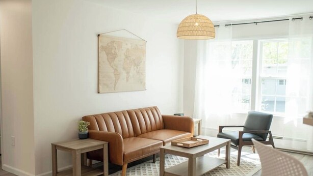 A modern living room with a brown leather sofa, wooden coffee table, and a small side table. A world map hangs on the wall, and a wicker pendant light is visible.