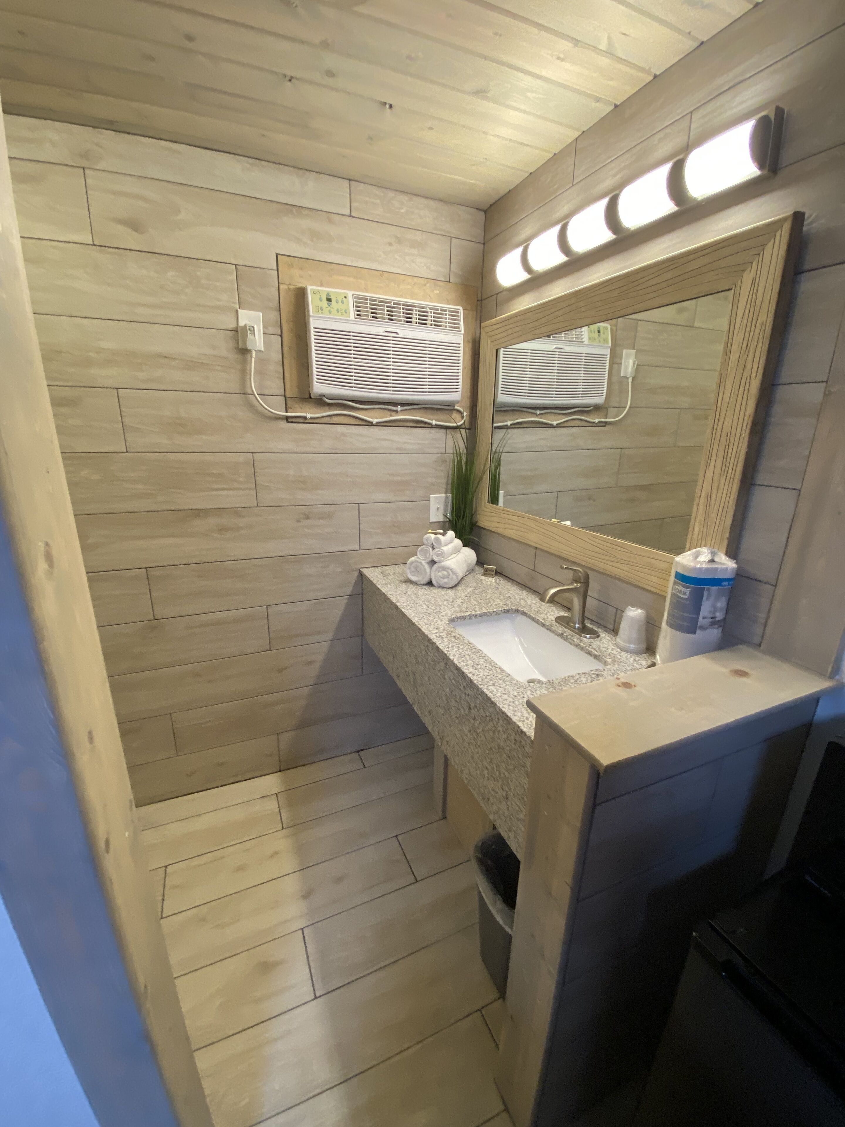 A bathroom with wooden walls, a granite countertop, a sink, a mirror, and a wall-mounted air conditioner.