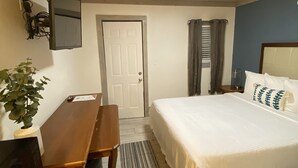 Ein Schlafzimmer mit Bett, Schreibtisch, Stuhl, einem an der Wand befestigten Fernseher und einer Tür.