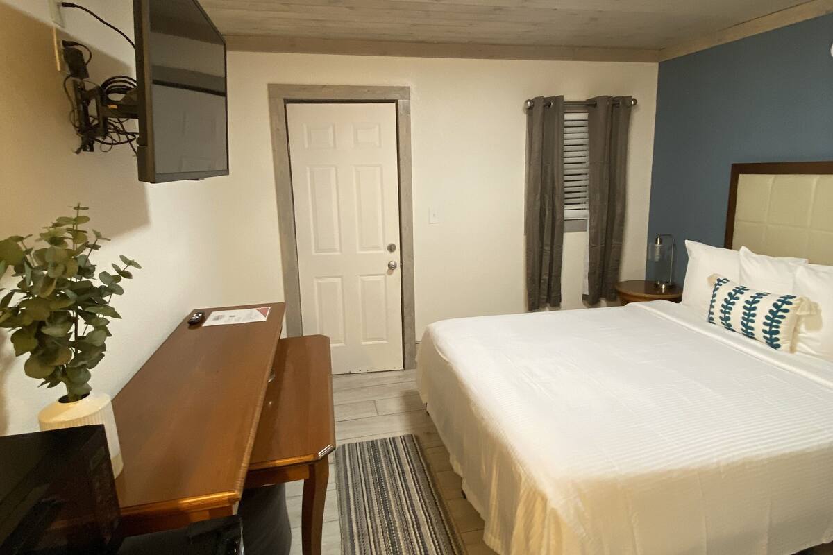 Ein Schlafzimmer mit Bett, Schreibtisch, Stuhl, einem an der Wand befestigten Fernseher und einer Tür.