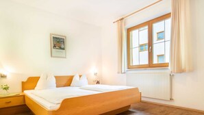A bedroom with a wooden bed, white bedding, a nightstand with a vase, a window with curtains, and a wall-mounted picture. - Apartment Neustift Near Stubai Ski Lift (Neustift Im Stubaital)