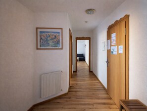 Un pasillo con suelo de madera, un radiador, un cuadro enmarcado en la pared y una puerta con avisos.