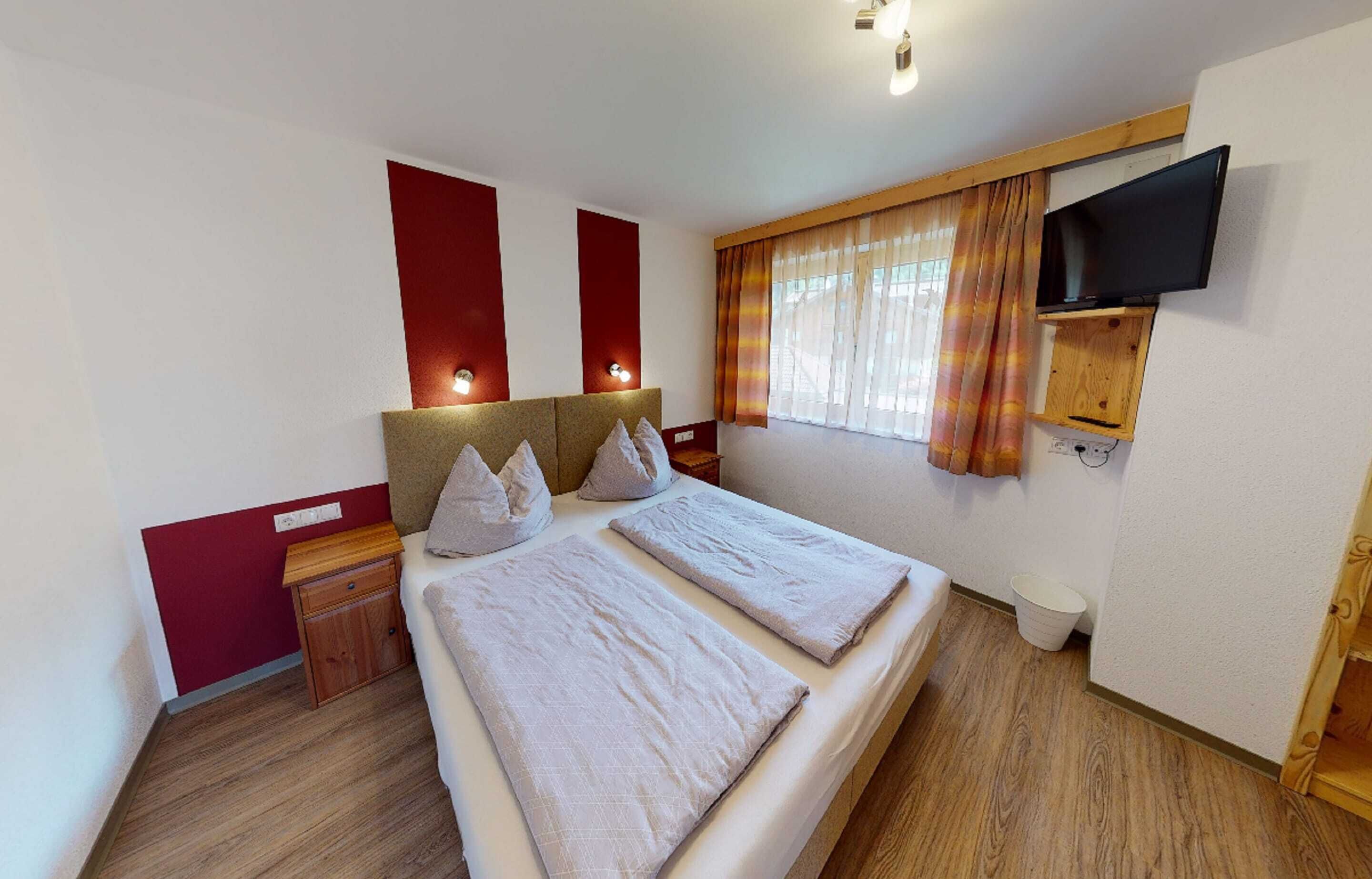 Un dormitorio con cama, mesitas de noche, televisión montada en la pared y ventana con cortinas.
