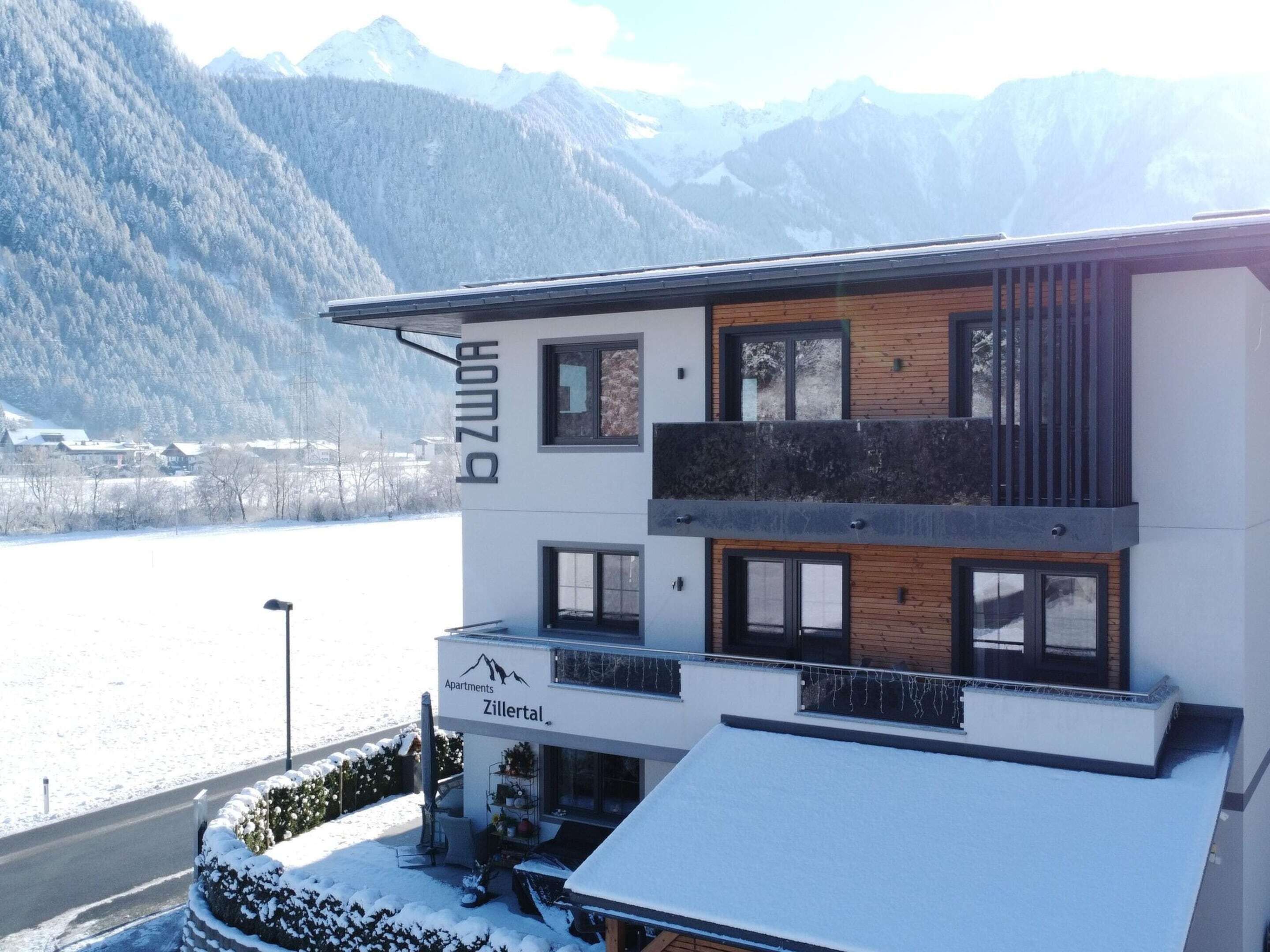 Un edificio moderno a due piani con un cartello che indica 'Zillertal Apartments' e vista su un paesaggio innevato con montagne sullo sfondo.