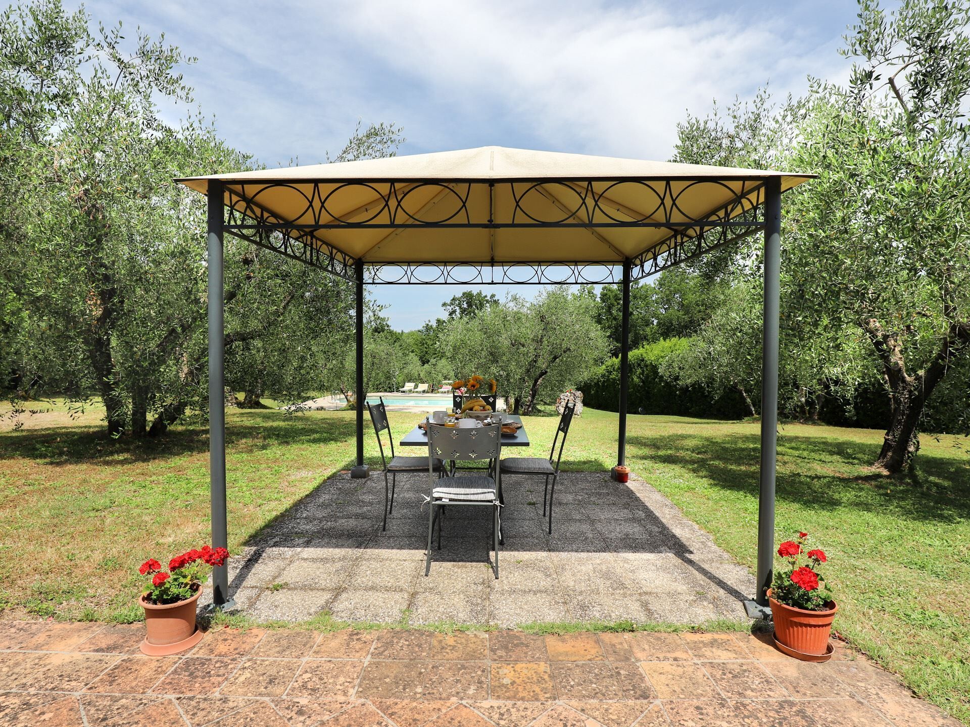 Un área de estar al aire libre con techo, con una mesa y sillas, rodeada de árboles y una piscina al fondo.