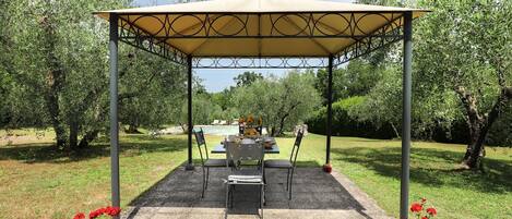 Un área de estar al aire libre con techo, con una mesa y sillas, rodeada de árboles y una piscina al fondo.
