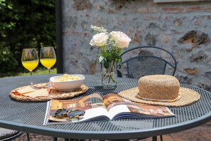 Una mesa metálica redonda con un mantel tejido, un plato de comida, un sombrero tejido, dos vasos de jugo de naranja, un jarrón con rosas blancas y una revista abierta.