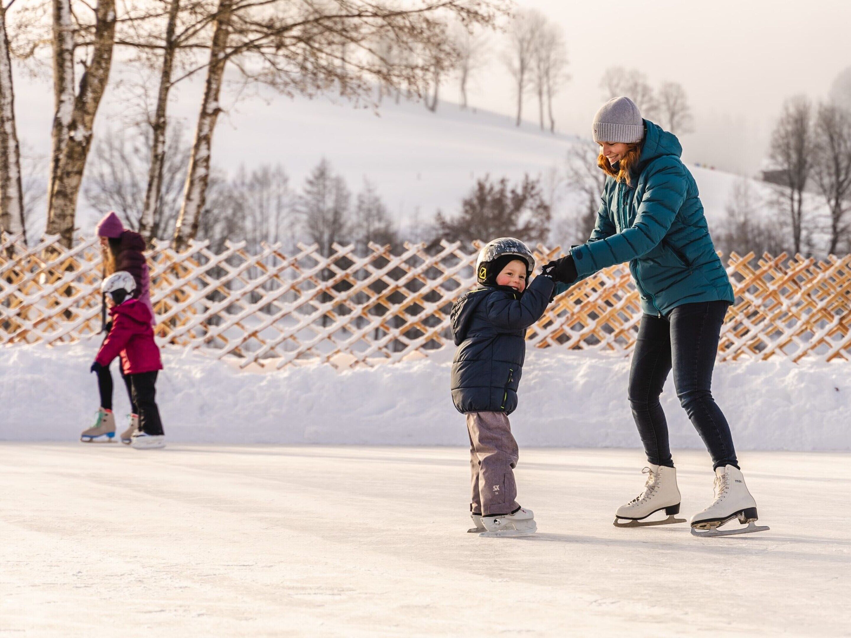 Una persona patina sobre hielo con un niño en una superficie helada, rodeados de nieve y árboles.