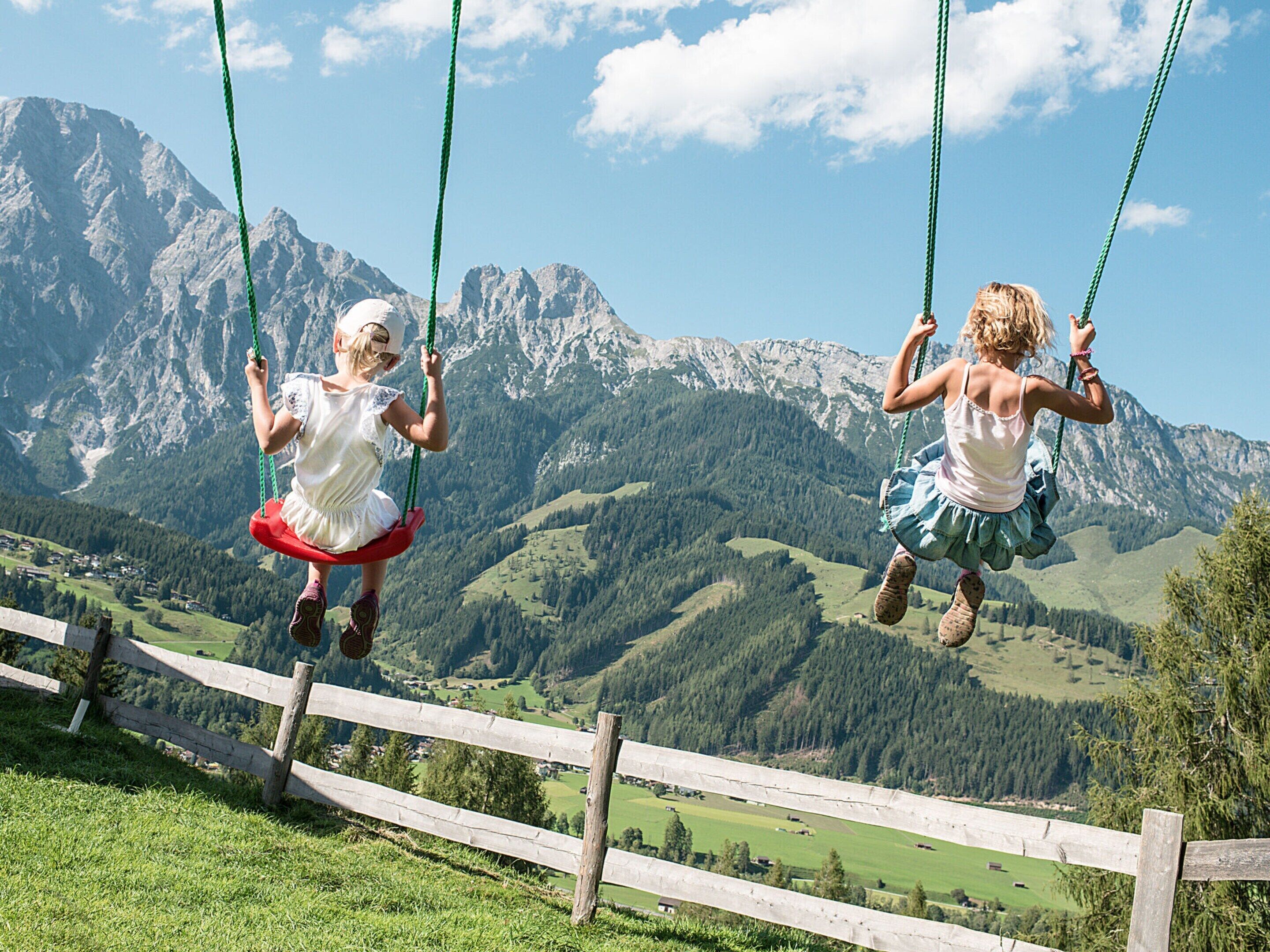 Two children on swings with a mountainous landscape in the background.