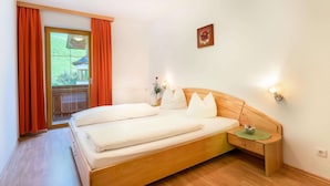 A bedroom with a wooden bed, orange curtains, a nightstand with a lamp, and a view of a house through a glass door. - Apartment in Stubaital Near Ski Lift (Neustift Im Stubaital)