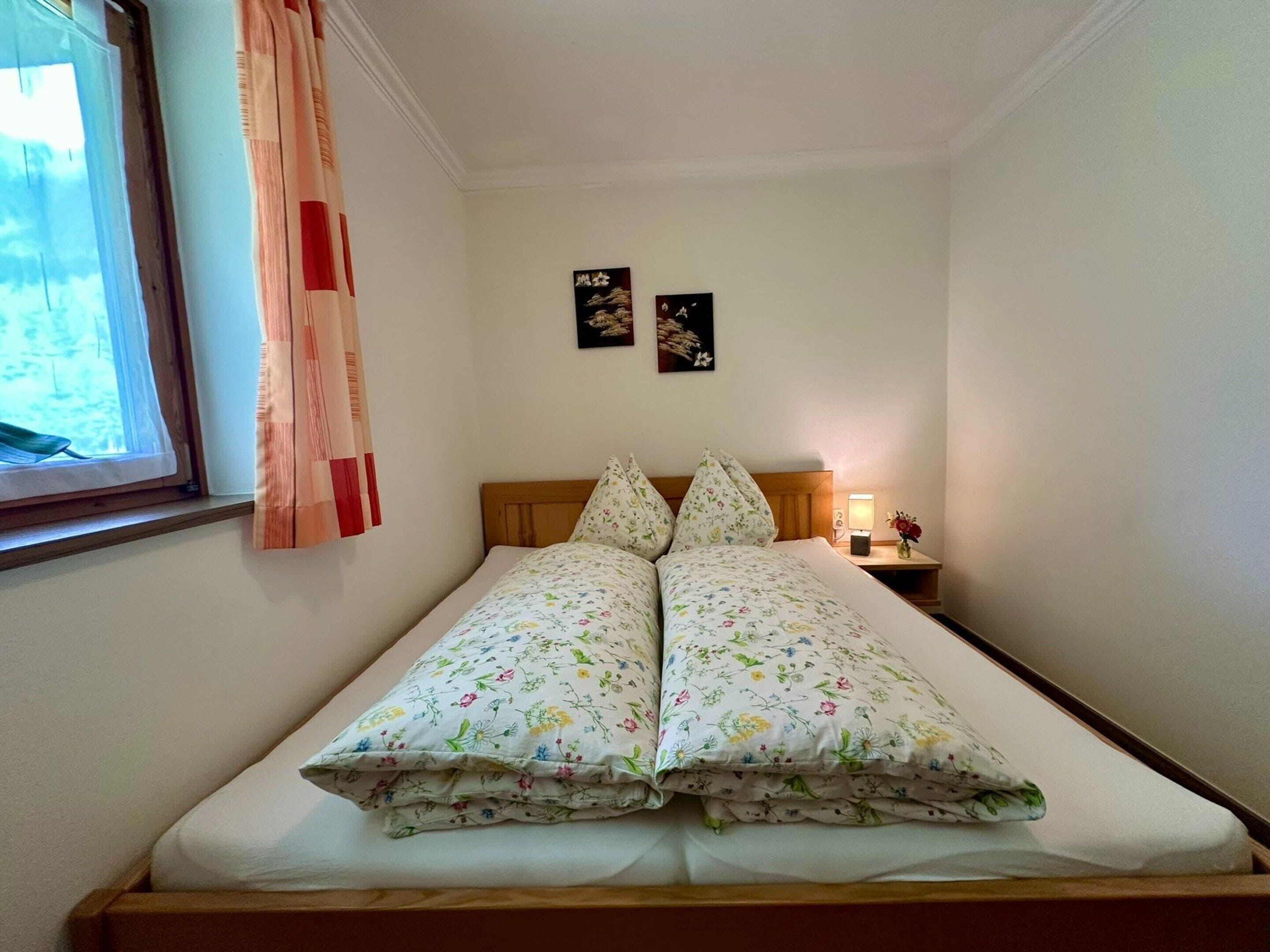 A single bed with floral bedding, a wooden headboard, a bedside table with a lamp, and two framed pictures on the wall.