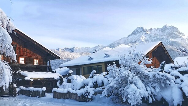 Une cabane en bois, dont le toit est recouvert de neige, avec une vue sur les montagnes en arrière-plan.