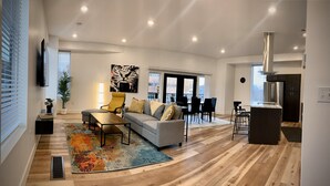 A modern living room with a sofa, armchairs, and a coffee table. There is a dining area with chairs and a table. The room has wooden flooring and large windows.