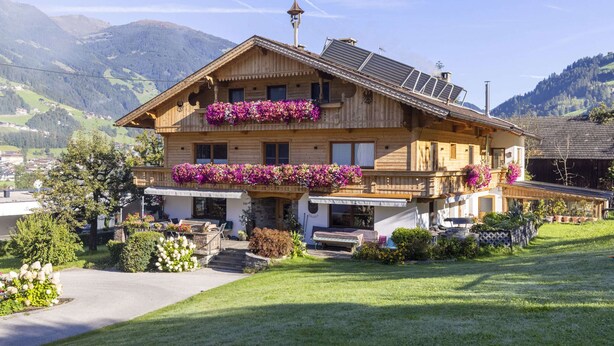 A wooden chalet with a balcony adorned with flowers, solar panels, and a stone chimney. - Moiklerhof Holiday Home in Ramsau im Zillertal (Ramsau im Zillertal)