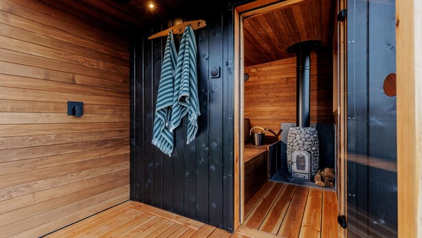 A sauna room with wooden walls and a glass door. There are towels hanging on a rack and a stone heater inside. - Lodge 19 (Wareham)