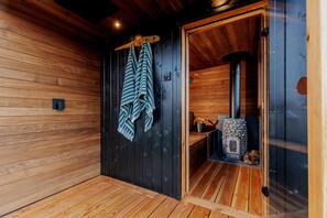 A sauna room with wooden walls and a glass door. There are towels hanging on a rack and a stone heater inside.