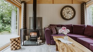 A cozy living room with a wooden ceiling, a large wall clock, a purple sofa, a wooden coffee table, and a log stack. - Lodge 19 (Wareham)
