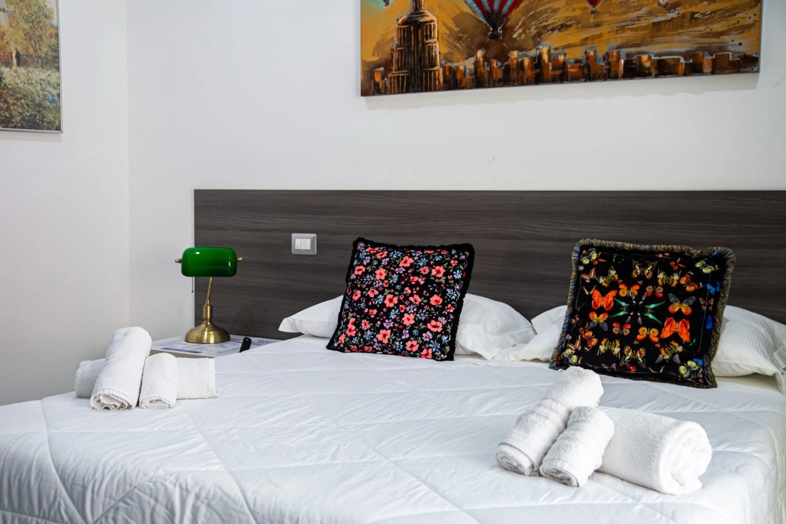 Una cama bien hecha con sábanas blancas, una lámpara de escritorio verde y dos almohadas decorativas con motivos florales.
