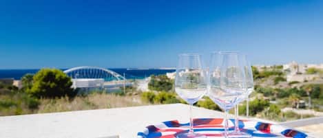 Two empty wine glasses on a colorful plate on a balcony with a clear view of the sea and a modern building in the background.
