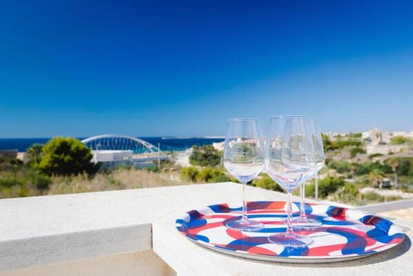 Two empty wine glasses on a colorful plate on a balcony with a clear view of the sea and a modern building in the background.