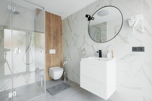 A modern bathroom with a glass-enclosed shower, a wooden accent wall, a white sink with a mirror, and a hairdryer on the wall.