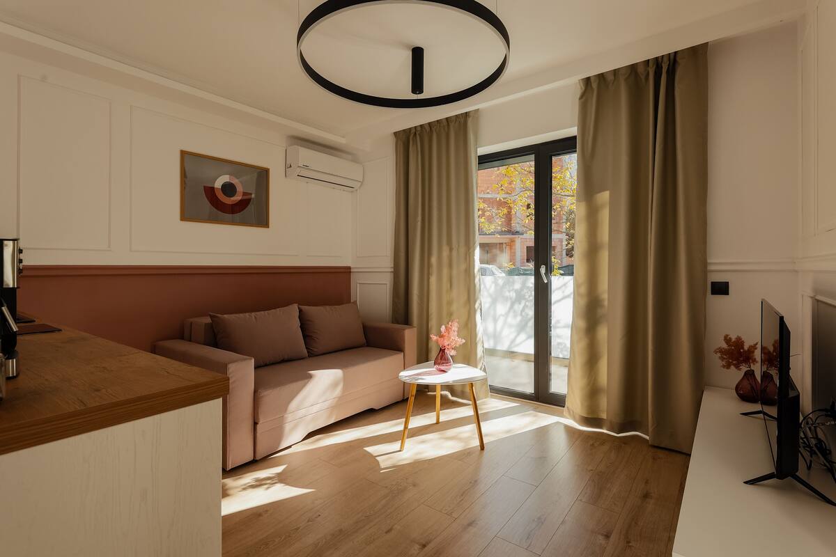 A modern living room with a sofa, a small table with a vase, a flat-screen TV, and a large window with curtains.