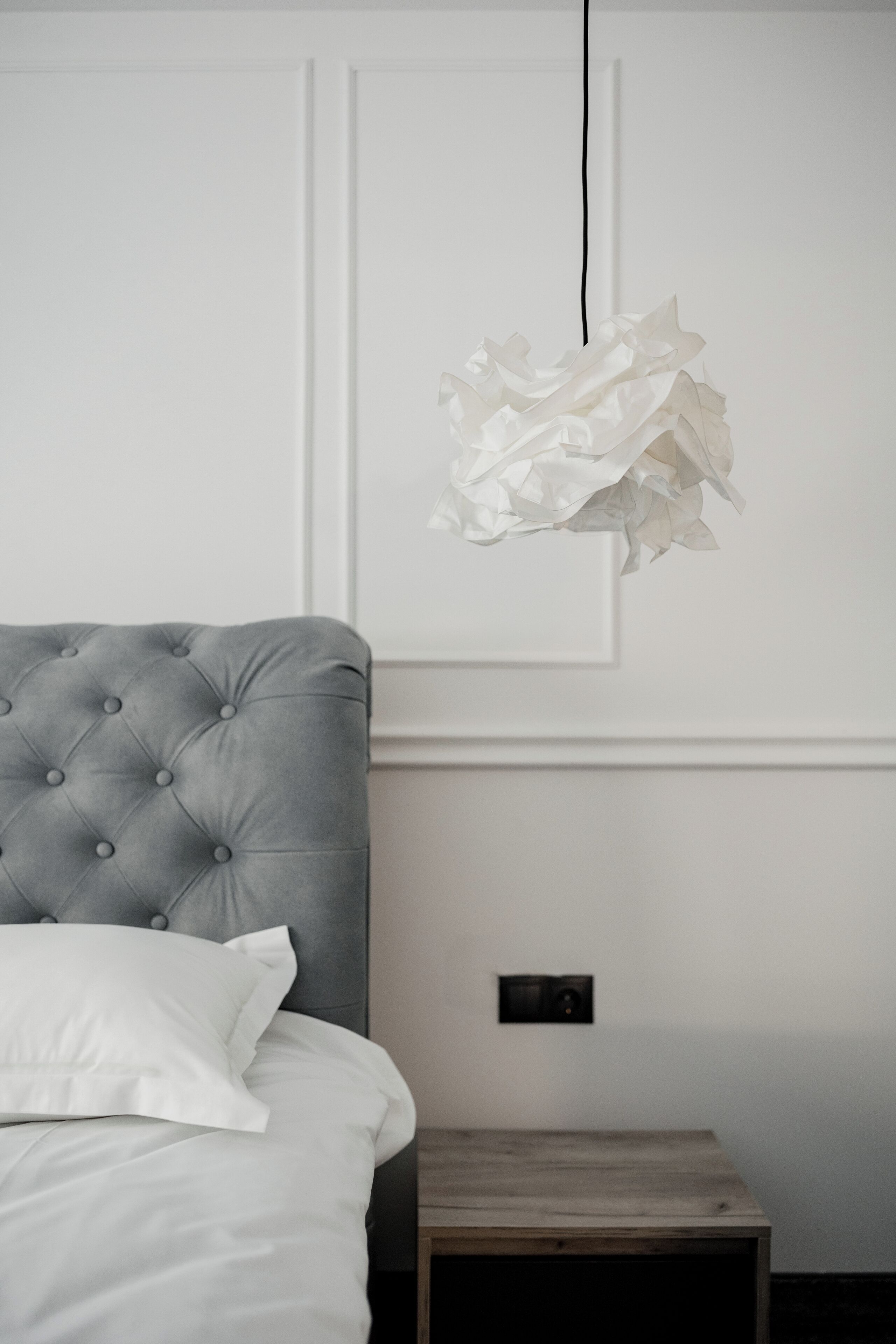 Un lit bien fait, avec une tête de lit grise et des oreillers blancs, une table de chevet et un luminaire fixé au mur.