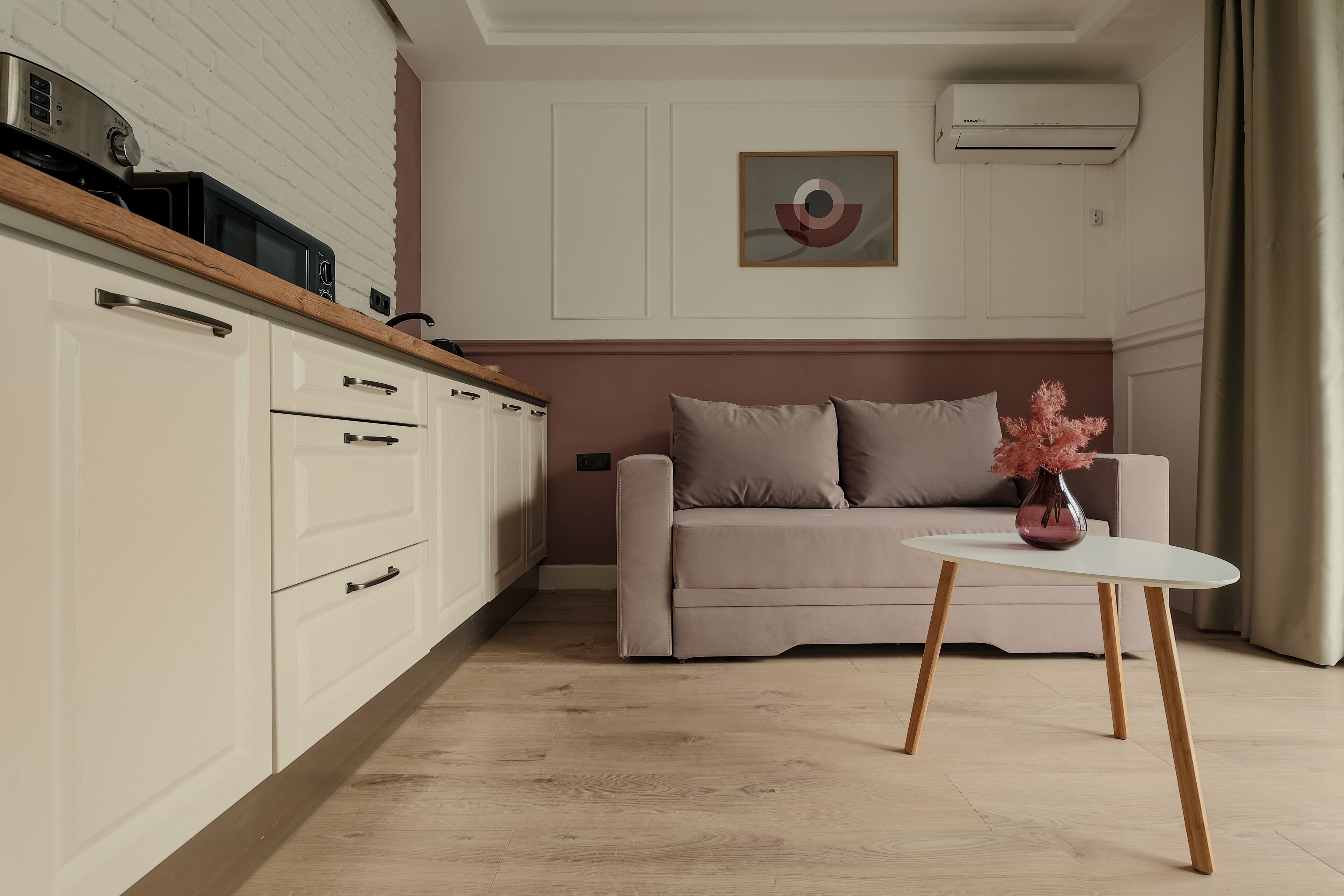 Une cuisine moderne avec des meubles blancs et un plan de travail en bois, un canapé beige agrémenté d’un vase de fleurs roses et une table blanche aux pieds en bois.