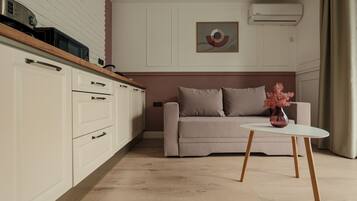 A modern kitchen with white cabinets and a wooden countertop, a beige sofa with a vase of pink flowers, and a white table with wooden legs.