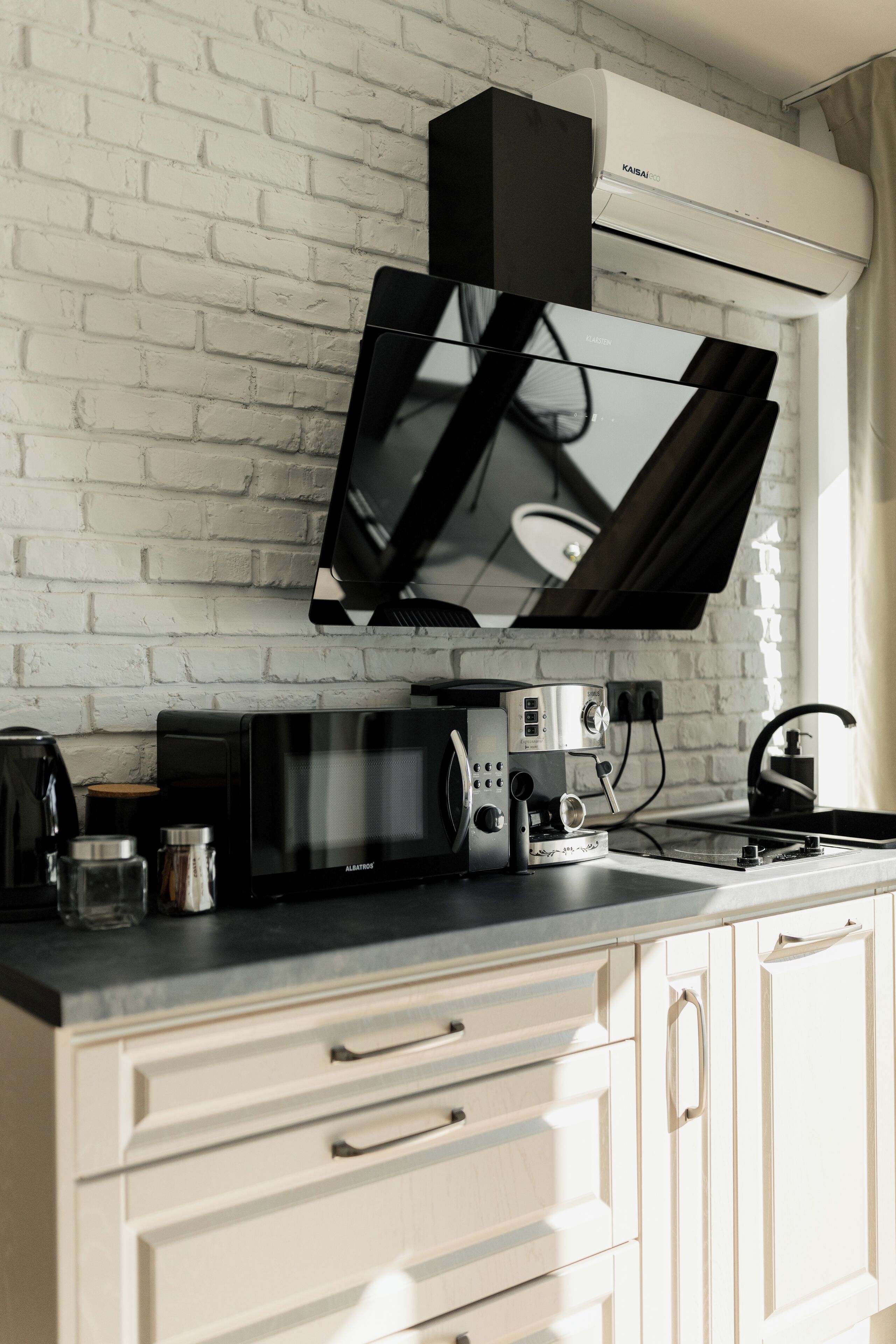 A modern kitchen with a white brick wall, a black range hood, a microwave, a coffee maker, and a sink.