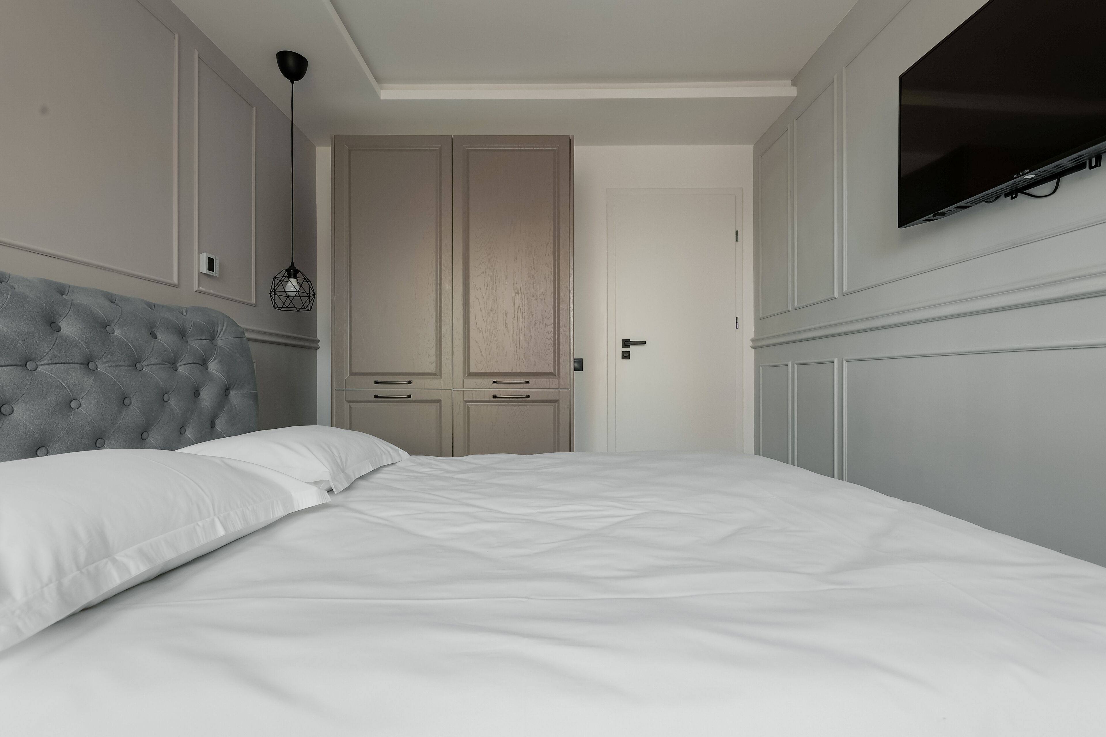 A bedroom with a tufted headboard, a bed with white linens, a wardrobe with a light wood finish, and a flat-screen TV mounted on the wall.
