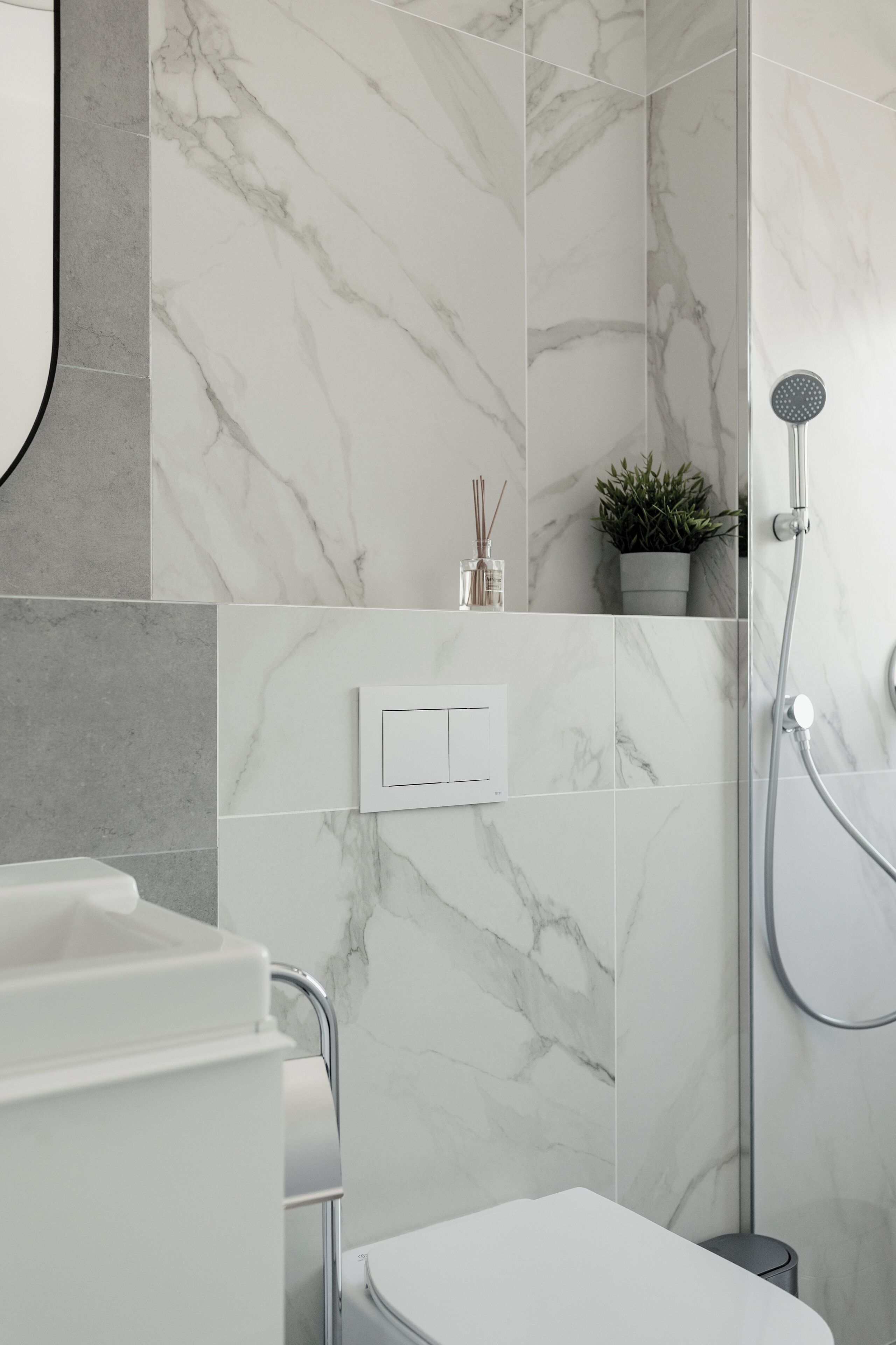 A modern bathroom with a shower, marble walls, a white sink, and a small plant on the shelf.