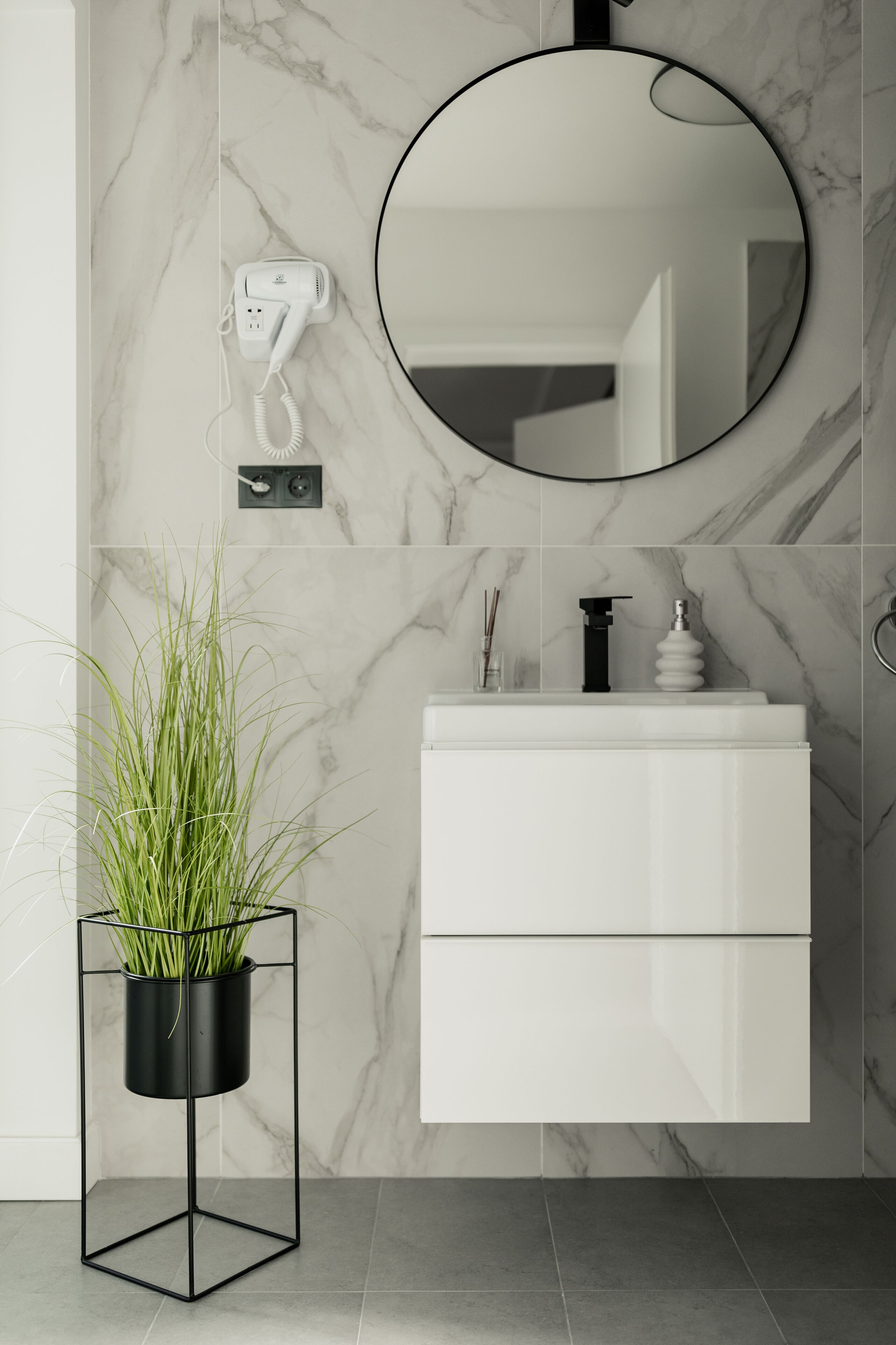A modern bathroom with a marble wall, a round mirror, a white vanity, a black plant in a metal stand, and a hairdryer on the wall.