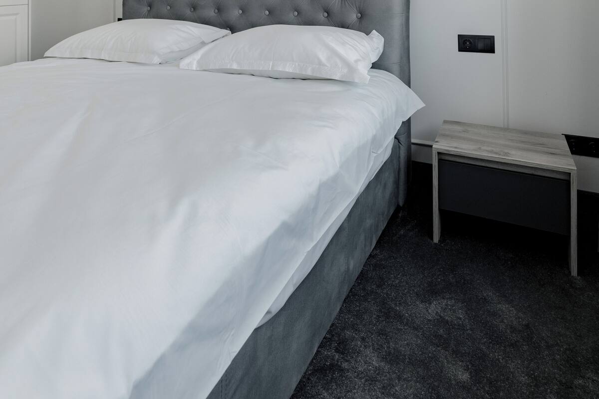 A neatly made bed with white linens, a tufted grey headboard, and a white wall paneling.