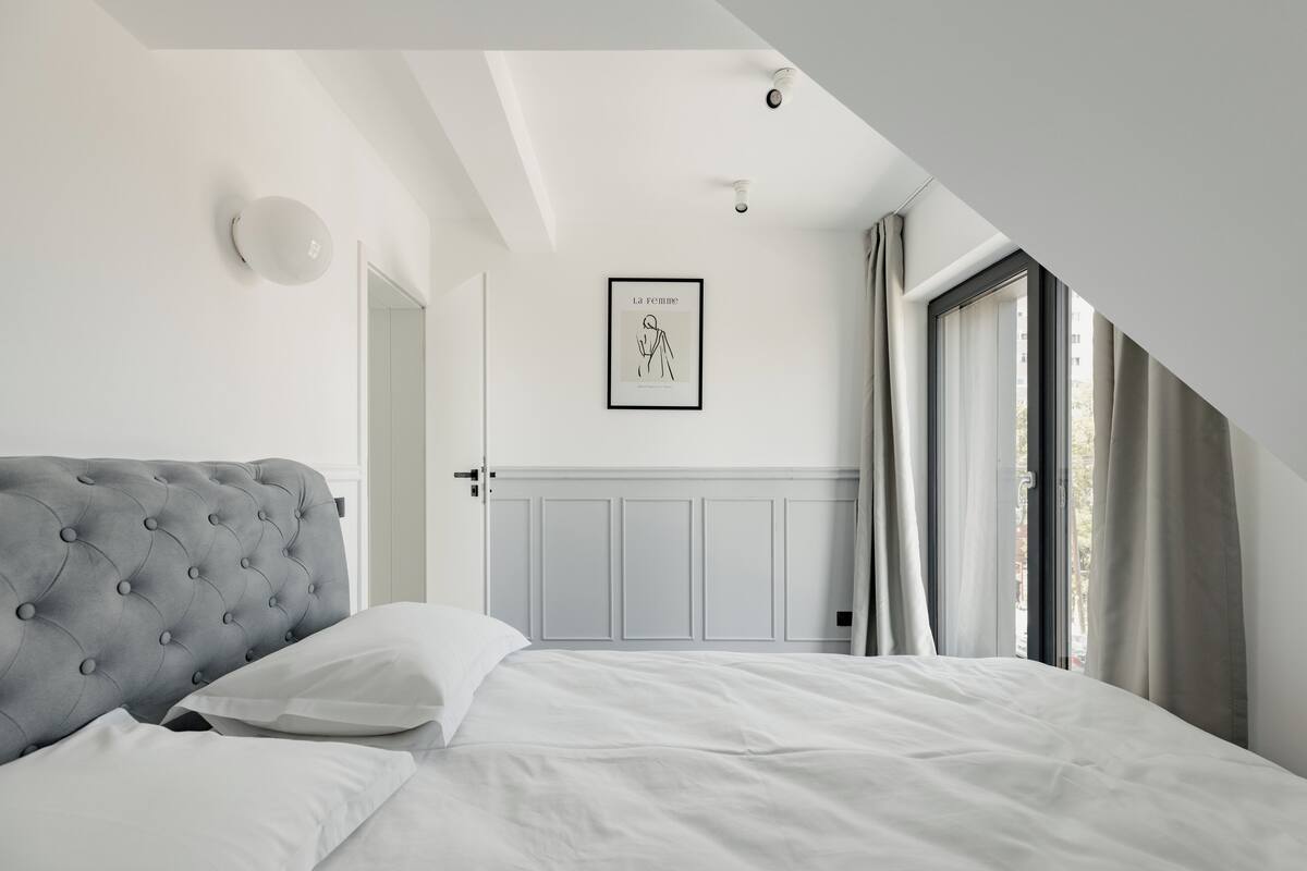 A bedroom with a tufted headboard, a framed artwork on the wall, and a window with curtains.