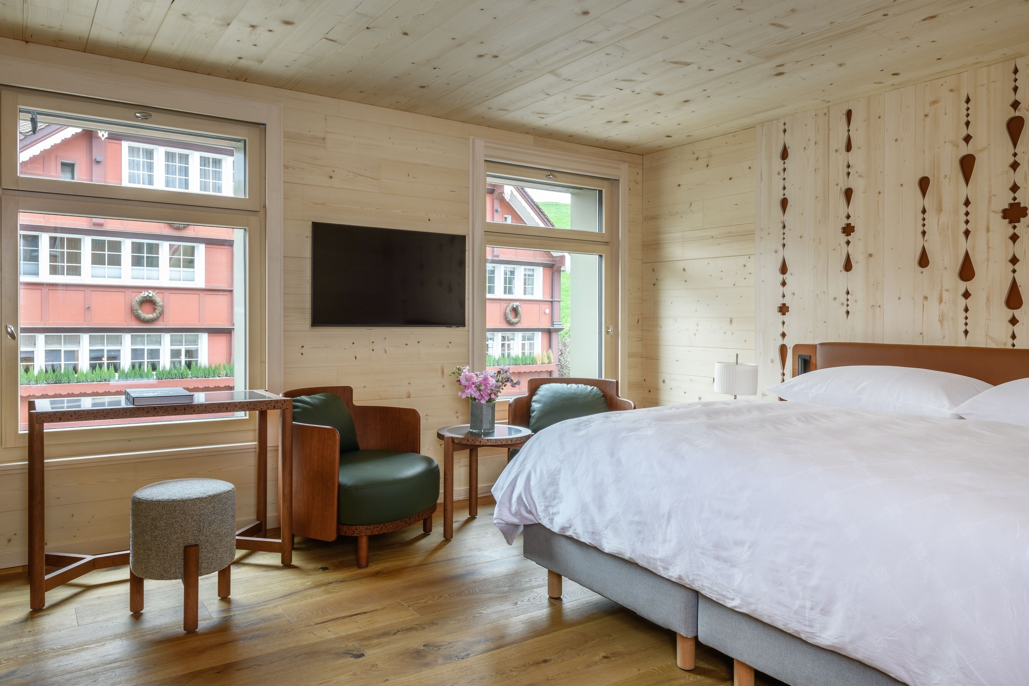 A hotel room with a bed, a desk, a chair, and a small stool. There are windows with views of buildings outside.