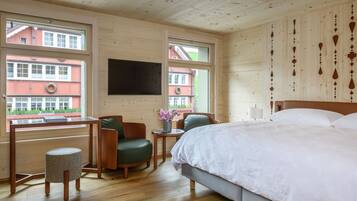 A hotel room with a bed, a desk, a chair, and a small stool. There are windows with views of buildings outside.