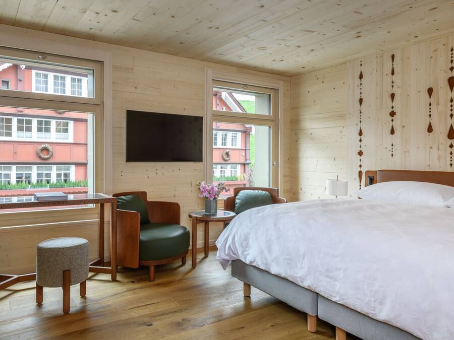 A hotel room with a bed, a desk, a chair, and a small stool. There are windows with views of buildings outside.