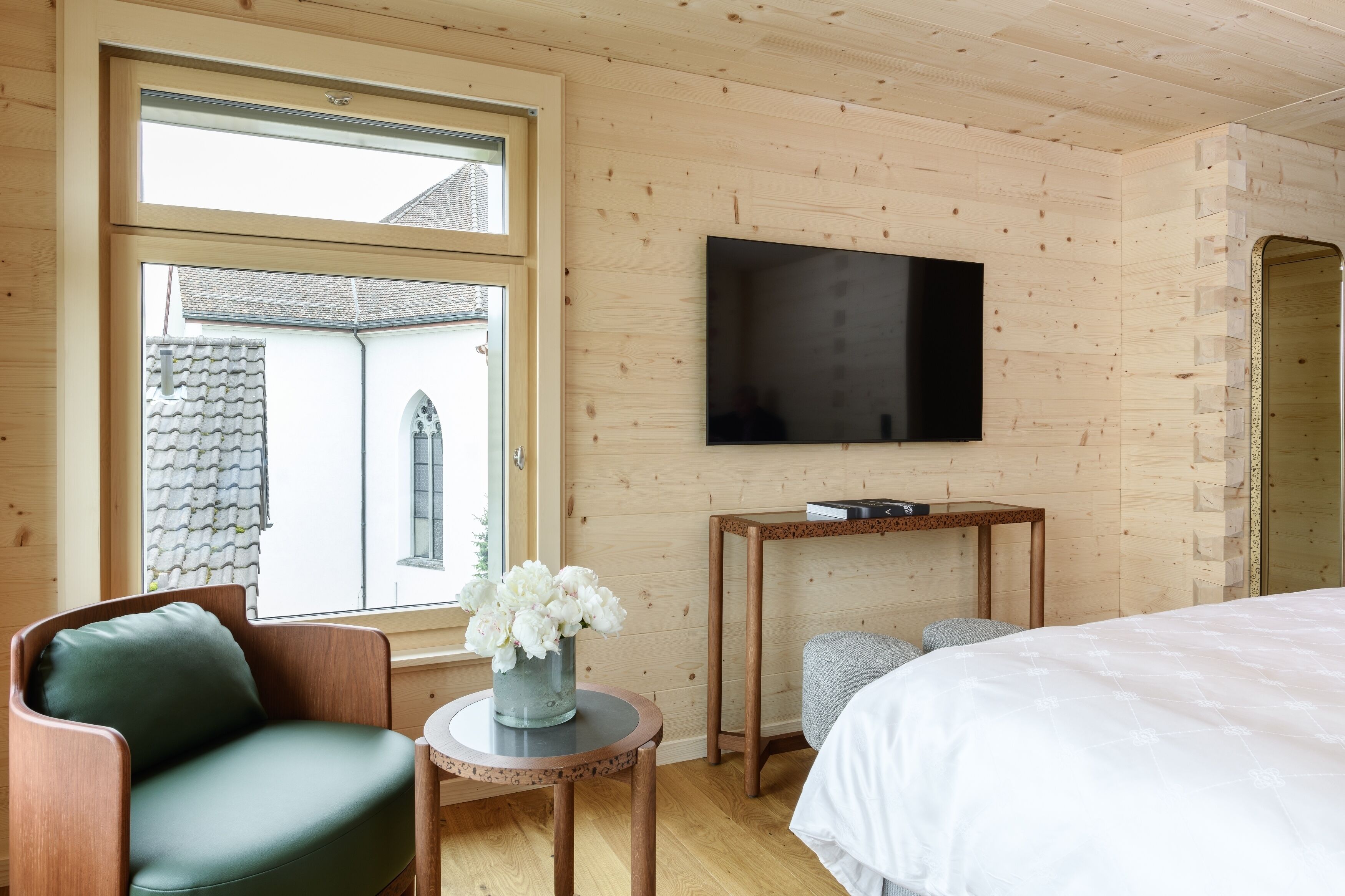 Ein Zimmer mit Holzvertäfelung, einem Bett, einem an der Wand befestigten Fernseher, einem Fenster mit Blick auf ein Gebäude und einem kleinen Tisch mit einer Blumenvase.