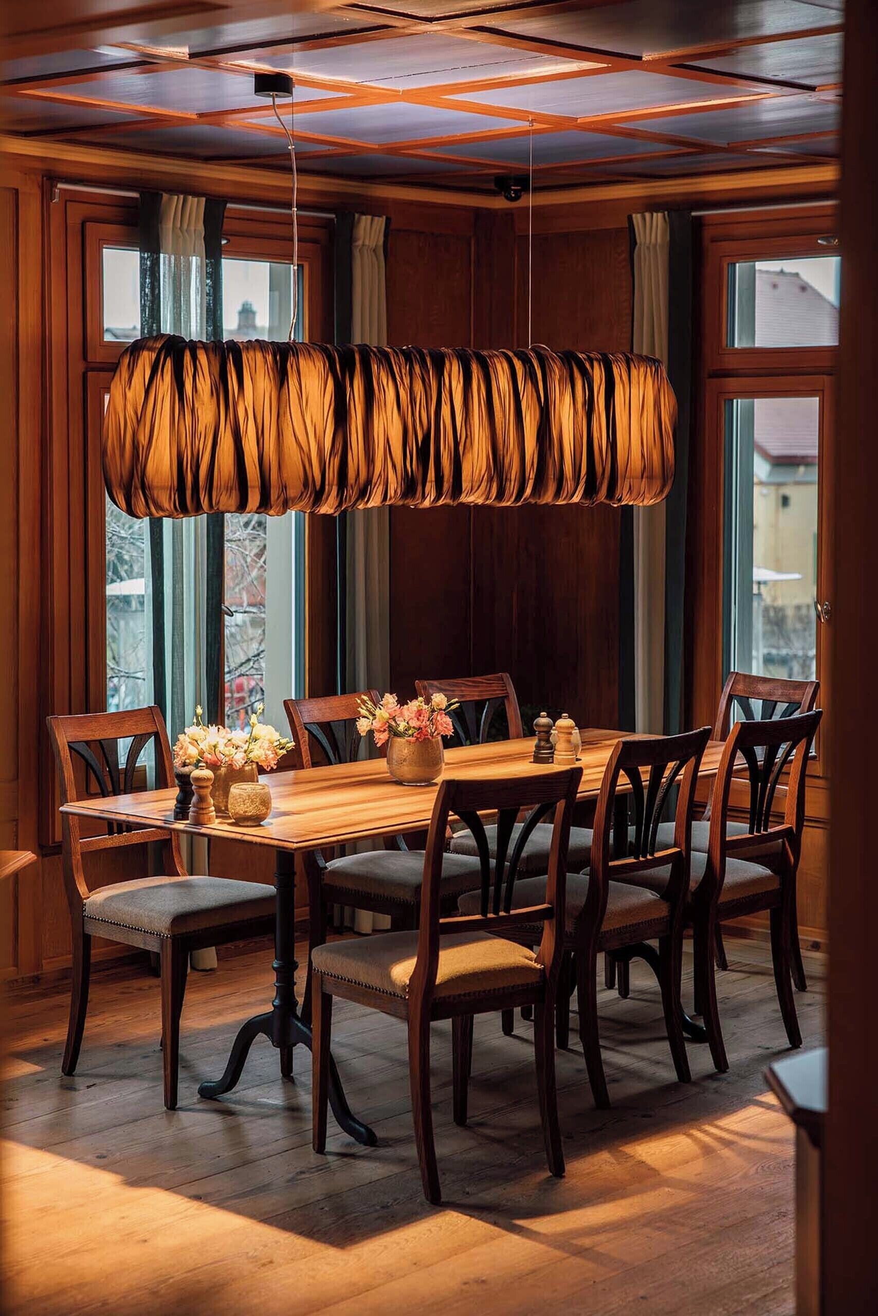 A dining area with a wooden table and chairs, a hanging light fixture, and large windows with curtains.