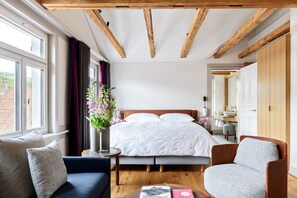 Une chambre avec un grand lit, un plafond en bois, une fenêtre avec des rideaux, une table de chevet ornée d’un vase de fleurs et une chaise.